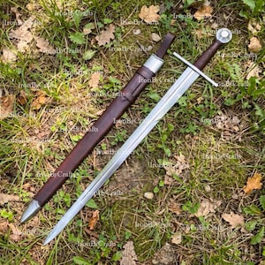 Puede incluir: Una espada de acero con una guarda recta y una empuñadura envuelta en cuero marrón oscuro, con un pomo redondo. A su lado yace una vaina de cuero marrón oscuro a juego con una contera y una boca de metal plateado, sobre la hierba.