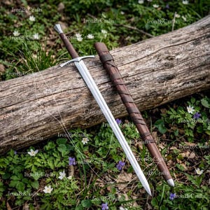 Peut inclure: Une épée en métal argenté avec une lame droite à double tranchant, une garde transversale simple et un manche marron foncé, accompagnée d'un fourreau en cuir marron foncé assorti enveloppé d'un cordon en cuir en spirale, posée sur une bûche.