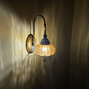 May include: A wall-mounted lamp featuring a curved brass-toned arm and a circular base plate. The light source is enclosed within a ribbed, amber-colored glass shade with a pumpkin-like shape, accented by a small decorative metal finial at the bottom.