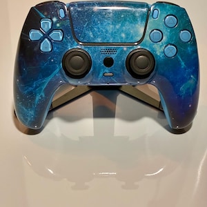 May include: A wireless video game controller featuring a glossy finish with a blue and black galaxy nebula print. The device includes two black analog sticks, a central touch pad, a directional pad, and four action buttons in light blue.
