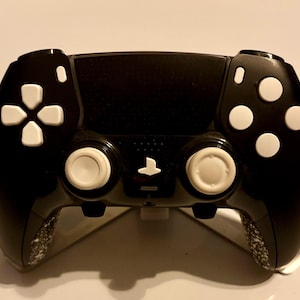 Puede incluir: Un mando de videojuegos negro con un panel táctil central, dos joysticks blancos, una cruceta blanca y cuatro botones de acción blancos. Las empuñaduras inferiores presentan un patrón moteado en blanco y negro y un logotipo blanco.
