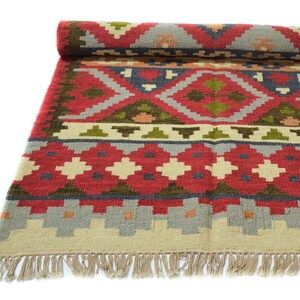 May include: A red, green and beige patterned rug with fringe. The rug has a geometric design with a central diamond shape.