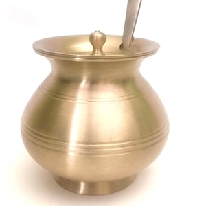 May include: A brushed brass sugar bowl with a rounded body, a wide flared rim, and a matching lid featuring a small central knob. A flat, brushed metal spoon is inserted into the opening of the lid, resting against the side of the bowl.