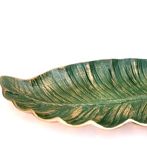 May include: A decorative tray shaped like a long, slender leaf. The surface features a deep green finish with textured gold-toned highlights along the veins and edges. The tray has a smooth, metallic-looking rim and a small stem at one end.