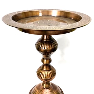 Puede incluir: Un soporte de pedestal decorativo de color bronce con una parte superior de bandeja redonda y poco profunda. La bandeja tiene un diseño floral grabado. El soporte tiene un diseño bulboso y escalonado con ranuras verticales. La base también es redonda con ranuras verticales.
