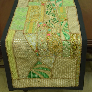 May include: A green and gold table runner with a patchwork design and beaded embellishments. The runner is made of various fabrics and has a black border.