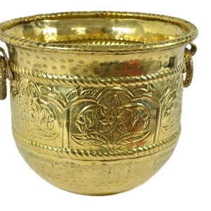 May include: A gold metal planter with a hammered finish and intricate floral designs. The planter has two handles on the sides.