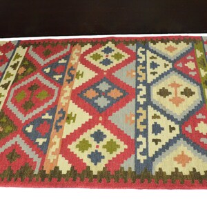 May include: A red, green, blue and beige patterned rug with fringe on the edges. The rug has a geometric design with diamond shapes and a tribal feel.