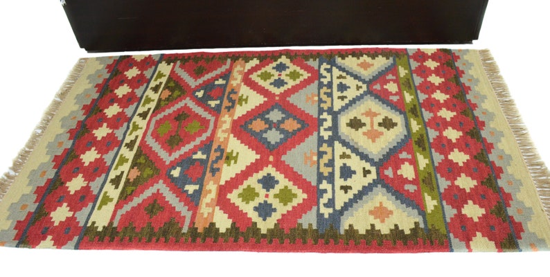 May include: A red, green, blue and beige patterned rug with fringe on the edges. The rug has a geometric design with diamond shapes and a tribal feel.