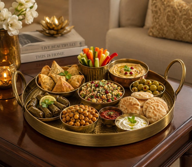 May include: A large round brass serving tray with two handles, holding several small brass bowls filled with various foods including samosas, stuffed grape leaves, roasted chickpeas, hummus, pita bread, olives, salad, and fresh vegetable sticks.