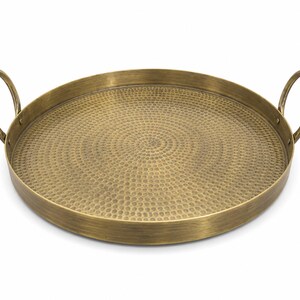 May include: A round brass tray with a brushed metallic finish and a textured hammered surface pattern. The tray features two circular handles attached to opposite sides of the raised rim, providing a functional design for carrying or display.