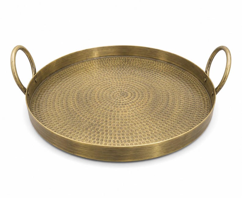 May include: A round brass tray with a brushed metallic finish and a textured hammered surface pattern. The tray features two circular handles attached to opposite sides of the raised rim, providing a functional design for carrying or display.