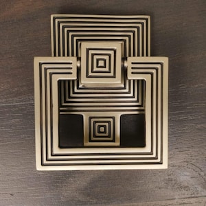 May include: A gold-toned metal cabinet pull with a geometric design. The pull has a square shape with multiple concentric squares inside.