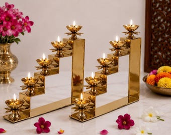 May include: Two gold-toned metal tiered candle holders, each featuring five stepped levels. Each level holds a small, lotus-shaped diya lamp with a lit flame. The structures are arranged side-by-side, creating a staircase effect with a polished finish.