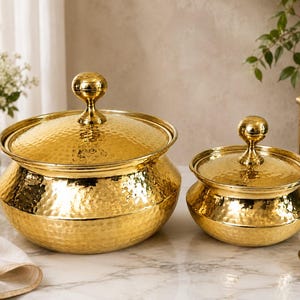 May include: Two round, gold-coloured metal serving bowls with matching lids. Each bowl features a hammered texture and a central knob handle on the lid. The bowls have a wide, rounded base that tapers towards the top rim, creating a distinct silhouette.
