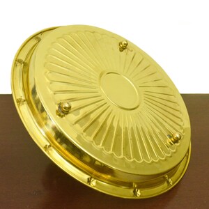 Puede incluir: Una bandeja de servir de metal dorado con un dise&ntilde;o decorativo de rayos de sol y cuatro peque&ntilde;os pies. La bandeja est&aacute; sobre una superficie de madera.