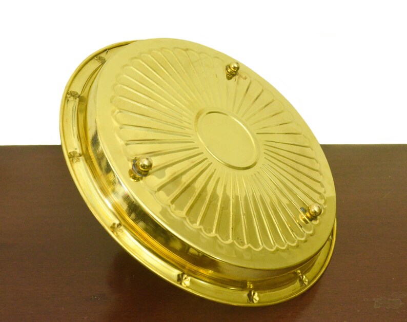 Puede incluir: Una bandeja de servir de metal dorado con un dise&ntilde;o decorativo de rayos de sol y cuatro peque&ntilde;os pies. La bandeja est&aacute; sobre una superficie de madera.