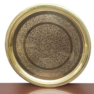May include: A round, brass-colored tray with an intricate floral pattern etched into the surface. The tray has a raised edge with a beaded border. The central design features a large flower surrounded by detailed foliage.