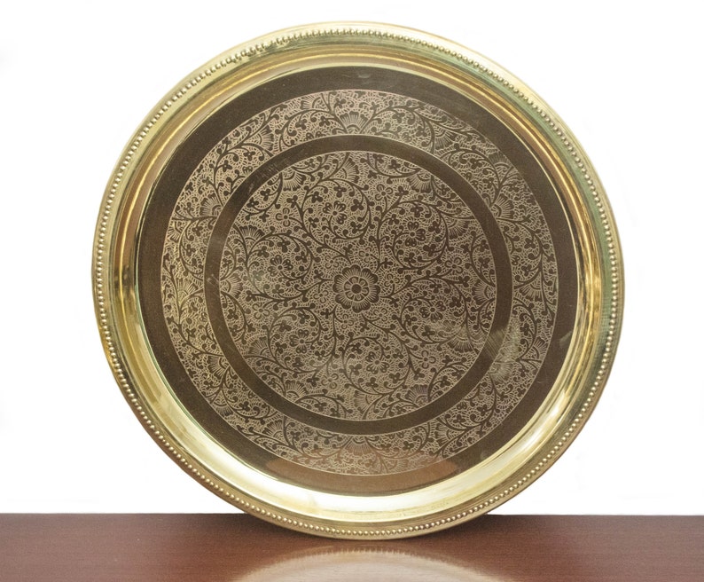 May include: A round, brass-colored tray with an intricate floral pattern etched into the surface. The tray has a raised edge with a beaded border. The central design features a large flower surrounded by detailed foliage.