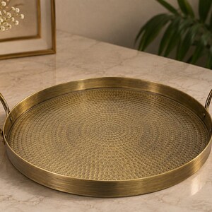May include: A round brass-colored metal tray featuring a textured, hammered surface pattern on the base. The tray includes two curved handles attached to opposite sides of the raised rim, providing a functional design for carrying or display.