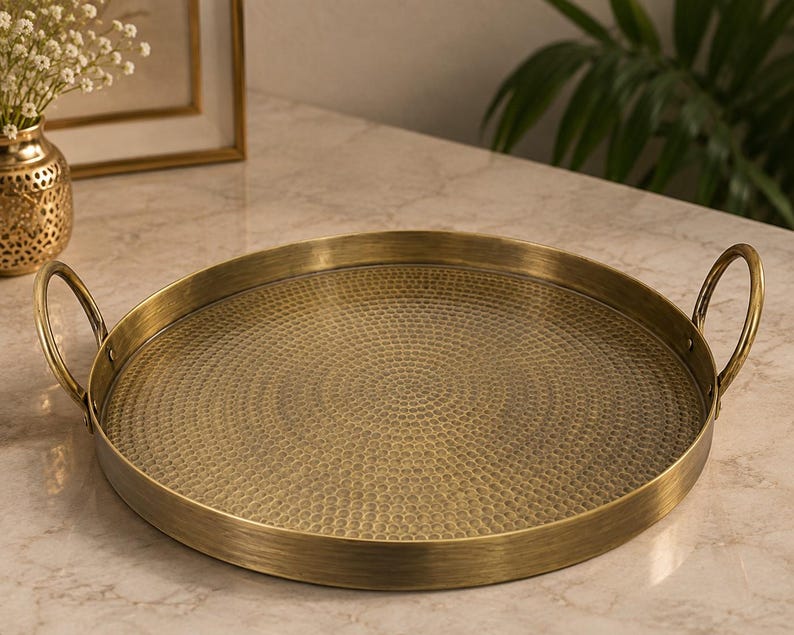 May include: A round brass-colored metal tray featuring a textured, hammered surface pattern on the base. The tray includes two curved handles attached to opposite sides of the raised rim, providing a functional design for carrying or display.