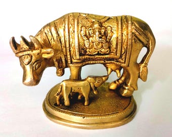May include: A gold-colored brass sculpture of a cow and calf. The cow is standing and has a decorative design on its back. The calf is standing next to the cow and is looking up at the cow. The sculpture is on a round base.