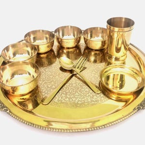 Op de afbeelding: Een goudkleurige metalen serveerset met een grote ronde schaal, kleine kommen, een hoog glas en een lepel en vork. De schaal en kommen hebben een decoratief patroon. De set is ontworpen voor het serveren van voedsel.