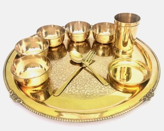 May include: A gold-colored metal serving set with a large round tray, small bowls, a tall glass, and a spoon and fork. The tray and bowls have a decorative pattern. The set is designed for serving food.