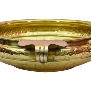 May include: A gold-colored metal bowl with an ornate design and a copper-colored handle. The bowl is round and has a wide, shallow base.