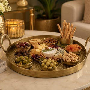 May include: A large, round, hammered brass serving tray with two arched handles, containing several small brass bowls filled with green olives, dark olives, grapes, dried apricots, pistachios, almonds, breadsticks, and a central cheese platter.