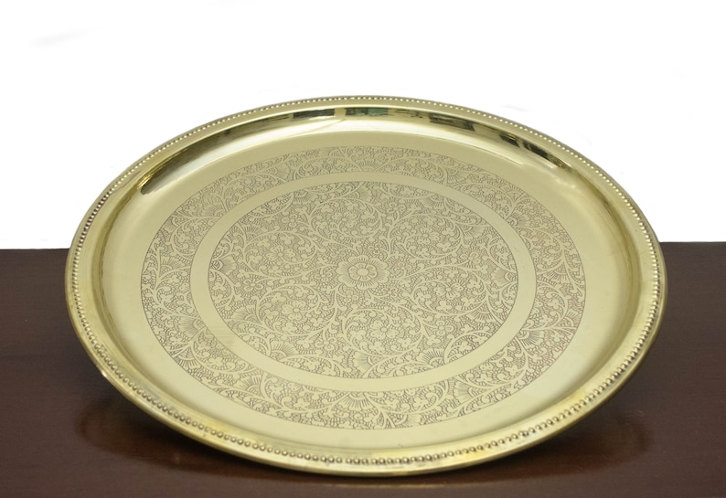 May include: A gold-colored, oval serving tray with an intricate floral pattern etched into the surface. The tray has a beaded edge and sits on a dark brown surface.