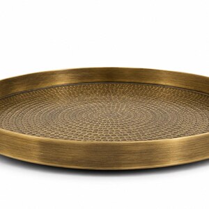 May include: A round brass-colored metal tray featuring a textured, hammered surface pattern on the interior base. The tray includes two curved, circular handles attached to opposite sides of the raised rim, finished with a brushed metallic sheen.