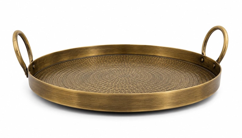 May include: A round brass-colored metal tray featuring a textured, hammered surface pattern on the interior base. The tray includes two curved, circular handles attached to opposite sides of the raised rim, finished with a brushed metallic sheen.
