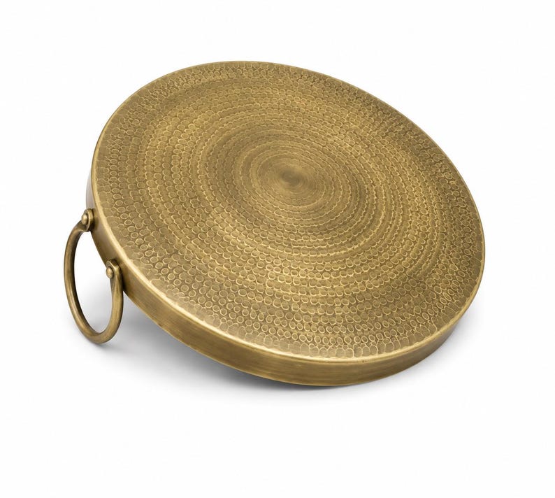 May include: A circular brass tray with a textured, hammered surface featuring concentric rings of small indentations. The tray has a smooth, raised edge and a single, round brass handle attached to one side, allowing for easy lifting and carrying.