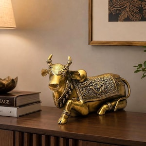 Peut inclure: Une figurine de vache dorée en position allongée, présentant des motifs gaufrés complexes sur la tête et une couverture décorative sur le dos avec un symbole Om proéminent. La sculpture possède des cornes, un collier et des bracelets détaillés.