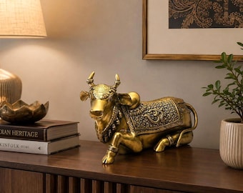 Könnte beinhalten: Eine goldfarbene Kuhfigur in liegender Position mit komplizierten Prägemustern am Kopf und einer dekorativen Decke auf dem Rücken, die ein markantes Om-Symbol zeigt. Die Skulptur hat detaillierte Hörner, eine Halskette und Beinschmuck.
