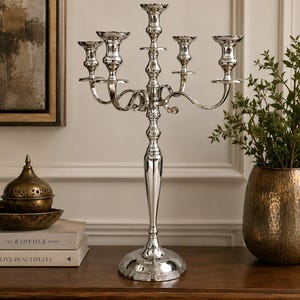 May include: A tall, polished silver-tone metal candelabra featuring a central pillar and four curved arms, each topped with a circular candle cup. The structure stands on a wide, tiered circular base with a smooth, reflective finish throughout.