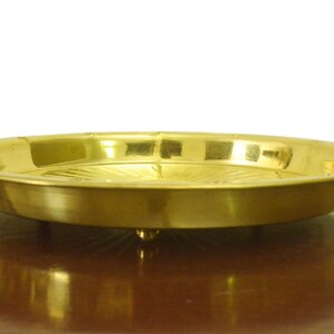 Puede incluir: Una bandeja redonda de servir de metal dorado con un borde elevado y un patr&oacute;n decorativo en la parte inferior. La bandeja est&aacute; sobre una superficie de madera.