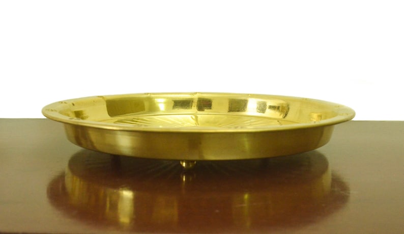 Puede incluir: Una bandeja redonda de servir de metal dorado con un borde elevado y un patr&oacute;n decorativo en la parte inferior. La bandeja est&aacute; sobre una superficie de madera.