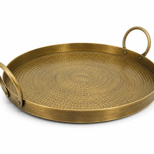 May include: A round brass serving tray with a hammered texture on the interior surface. The tray features a smooth, raised rim and two curved metal handles attached to opposite sides with visible rivets, providing a functional grip.