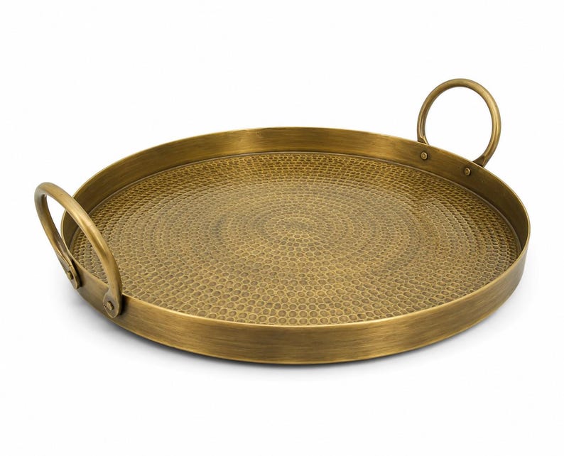 May include: A round brass serving tray with a hammered texture on the interior surface. The tray features a smooth, raised rim and two curved metal handles attached to opposite sides with visible rivets, providing a functional grip.