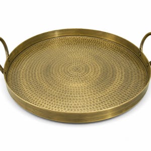 May include: A round brass tray with a hammered texture on the interior surface. The tray features a raised rim and two curved handles attached to opposite sides. The metal has a brushed, antique gold finish with a consistent circular pattern.