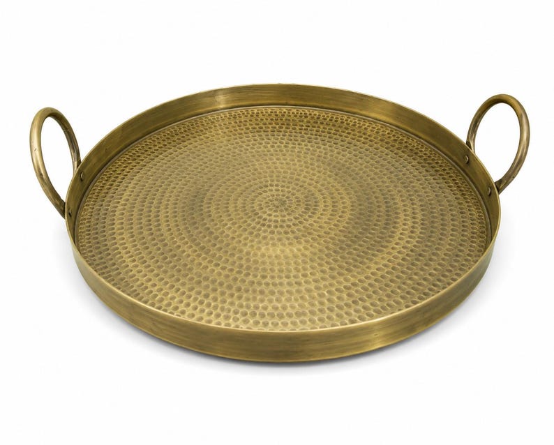 May include: A round brass tray with a hammered texture on the interior surface. The tray features a raised rim and two curved handles attached to opposite sides. The metal has a brushed, antique gold finish with a consistent circular pattern.