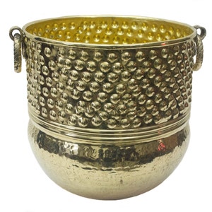 Puede incluir: Un macetero grande de metal dorado con dos asas. El macetero tiene una textura martillada y una banda de puntos en relieve alrededor del centro.