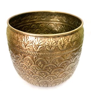 May include: A gold-colored metal planter with intricate embossed floral and geometric patterns. The planter has a wide, open top and a rounded body. The detailed design covers the entire surface, creating a textured appearance. The planter is suitable for indoor or outdoor use.