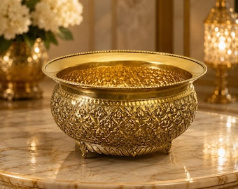 May include: A polished gold-colored metal bowl featuring an intricate embossed floral pattern across its exterior surface. The bowl has a wide, smooth rim, a rounded body, and rests on small, decorative feet, showcasing a reflective metallic finish.