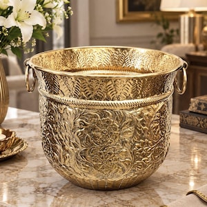 May include: A polished gold-tone metal bucket featuring an intricate embossed floral pattern across the exterior, a textured rope-style band encircling the upper section, and two circular side handles attached to the rim of the vessel.
