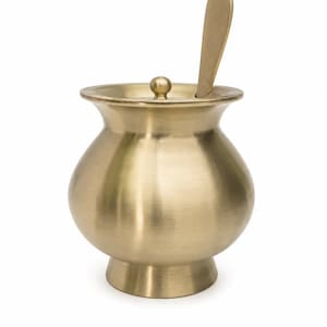Puede incluir: Un recipiente de latón cepillado con cuerpo redondeado, borde acampanado y base de pedestal. El recipiente incluye una tapa circular a juego con un pequeño pomo central y una cuchara plana de latón que descansa dentro de la abertura.