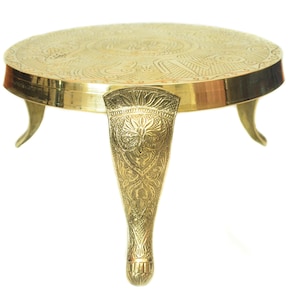 May include: A gold metal decorative stand with a round top and three legs. The legs are intricately carved with floral designs.