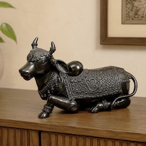 Peut inclure: Une statue en bronze foncé représentant un taureau couché. Le taureau présente des motifs en relief complexes sur le corps, une couverture décorative drapée sur le dos avec un symbole Om, et des bijoux ornés au cou et aux pattes avec des cornes.